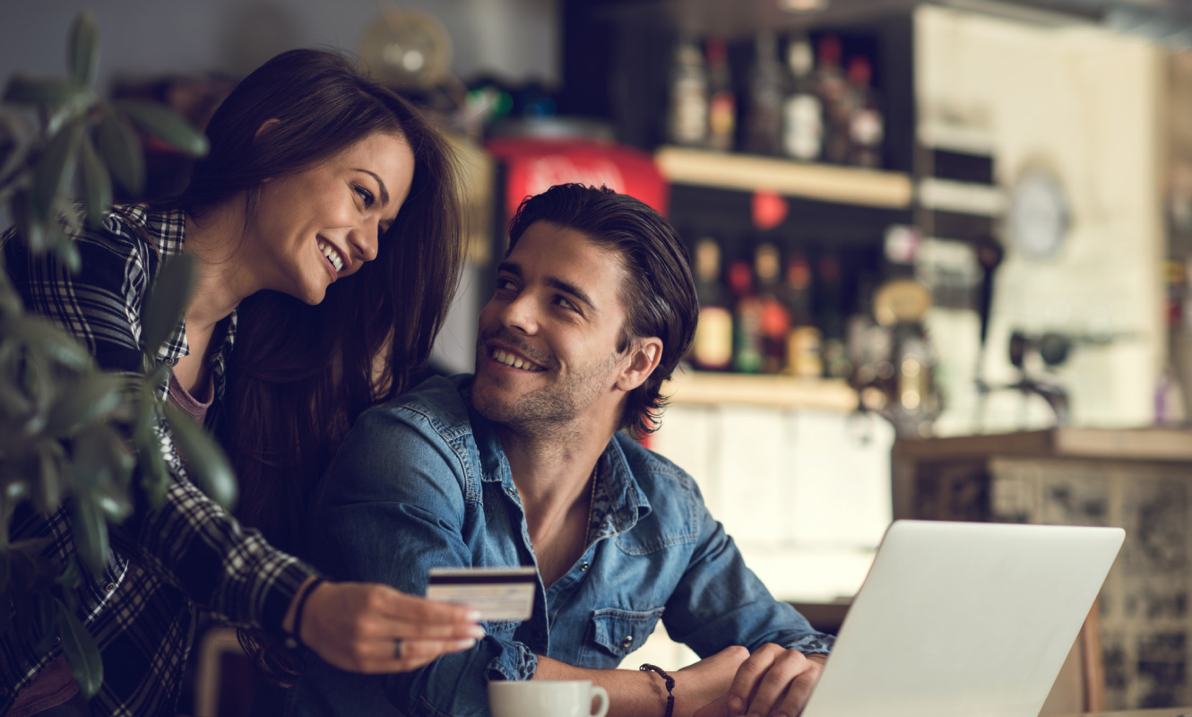 A man and woman in front of a laptop. The woman is holding a credit card. They are both smiling.