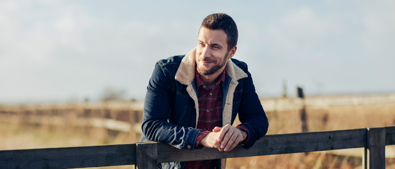 A bearded man in a denim jacket leaning against a wooden fence.
