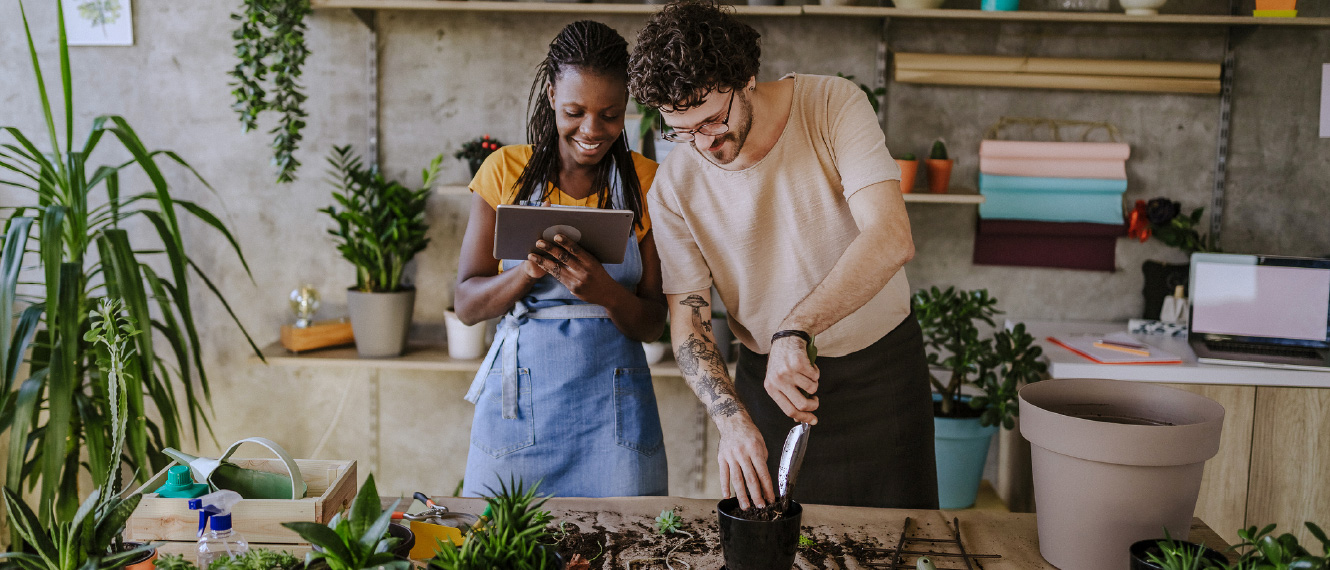 Two young adults potting plants at a large table. The woman is holding a tablet.