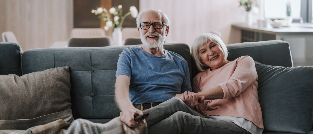 An older couple relaxing on a couch, both are smiling at the camera.