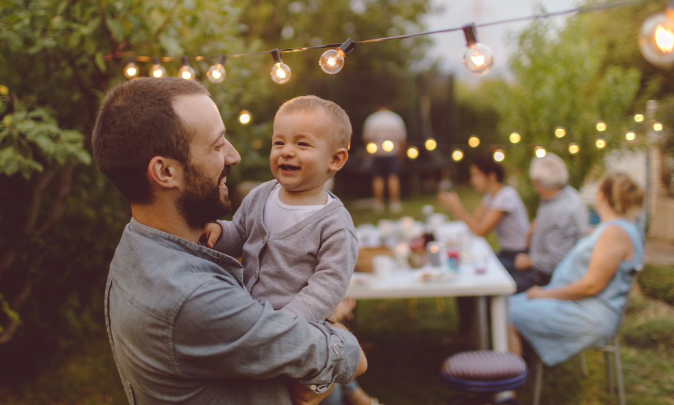 A young man holding a toddler, they are both smiling. There's a family gathering going on behind them.